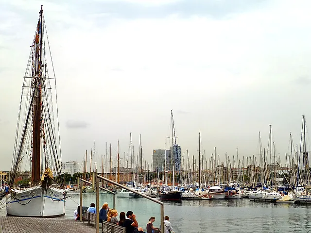 Barche a vela ormeggiate nel porto turistico del Port Vell, con un alto albero di legno in primo piano